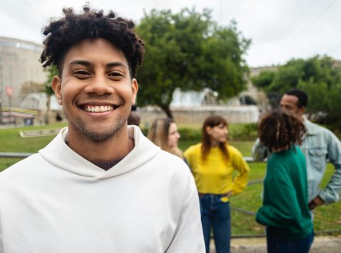 Smiling hispanic latin young man looking at camera outdoors. Portrait of african american millennial guy standing outdoors with his friends on the background. Friendship, happiness and youth concept.