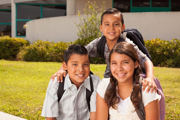 Young Hispanic Student Children Wearing Backpacks On School Campus.
